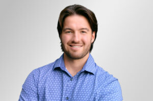 A photo of a man with brown hair and a blue dress shirt. He is smiling